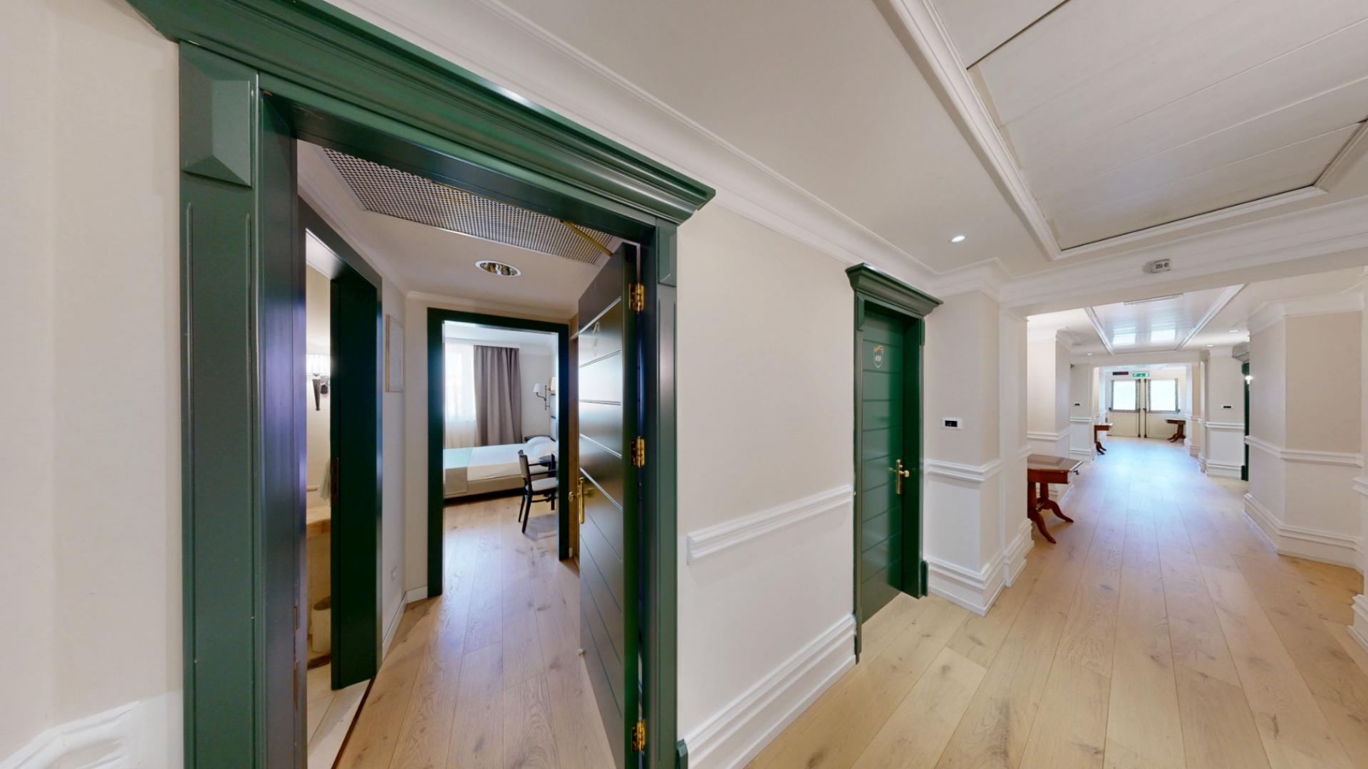 Corridoio elegante con porte verdi, pavimento in legno e una stanza visibile.