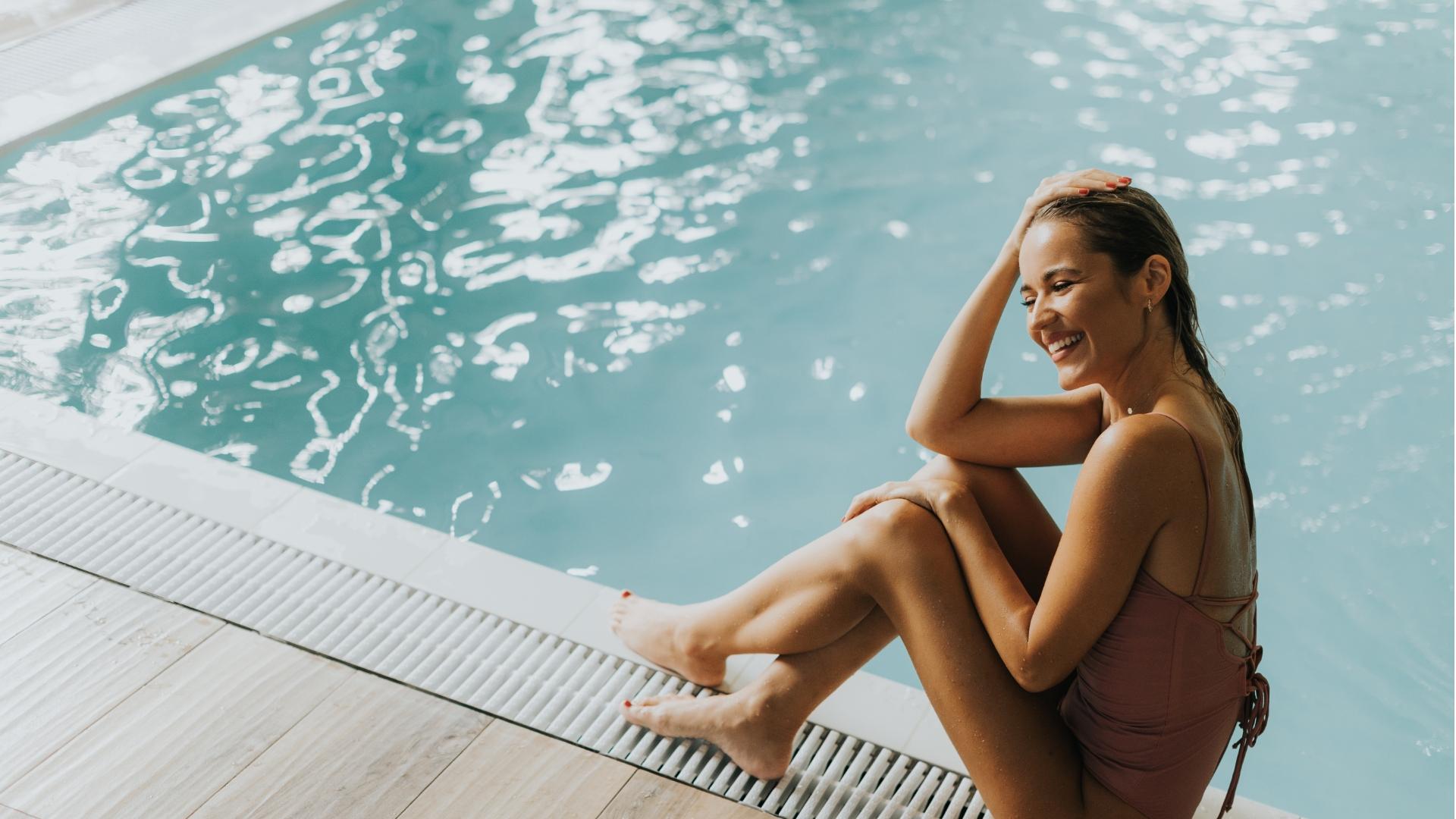 Donna sorridente seduta a bordo piscina con costume intero rosa.