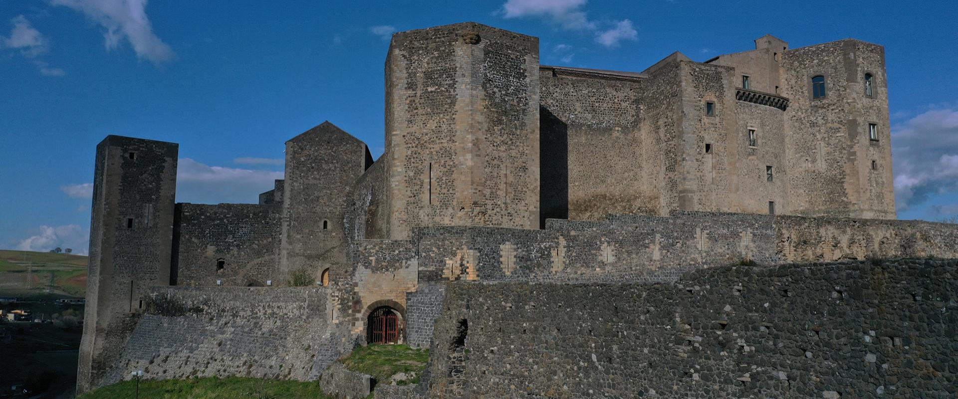 Castello medievale su collina verde, circondato da montagne e cielo azzurro.