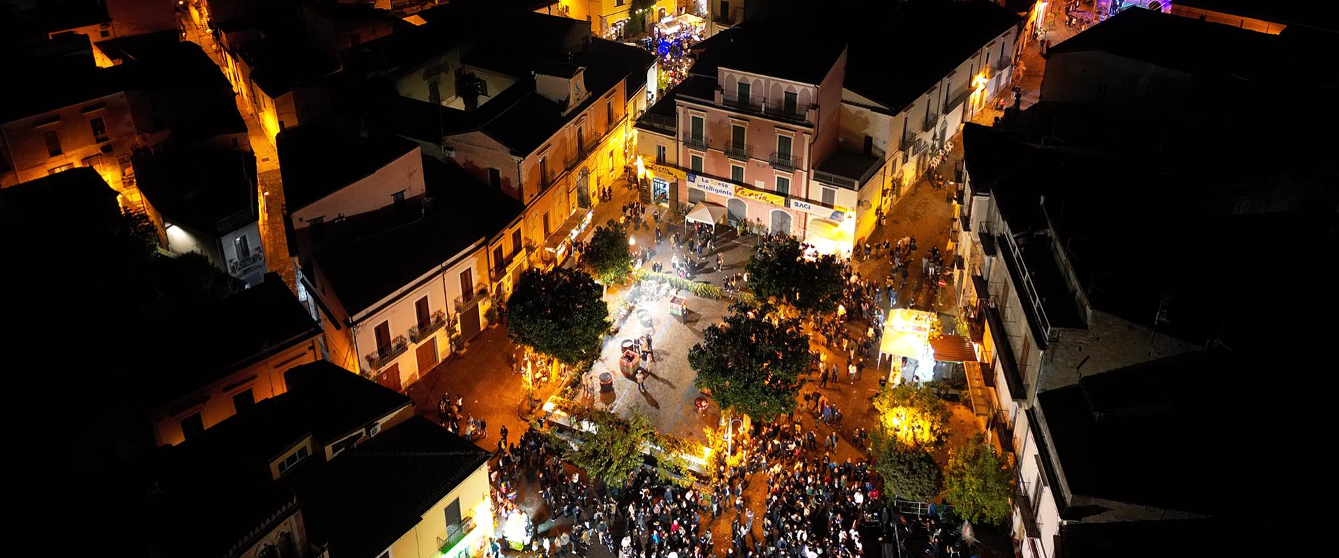 Vista notturna di una città illuminata, folla festosa riempie una piazza vibrante di vita.