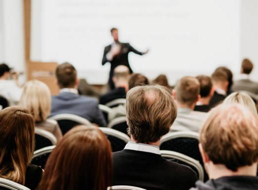 Conferenza con partecipanti seduti, relatore sul palco, atmosfera professionale e formale.