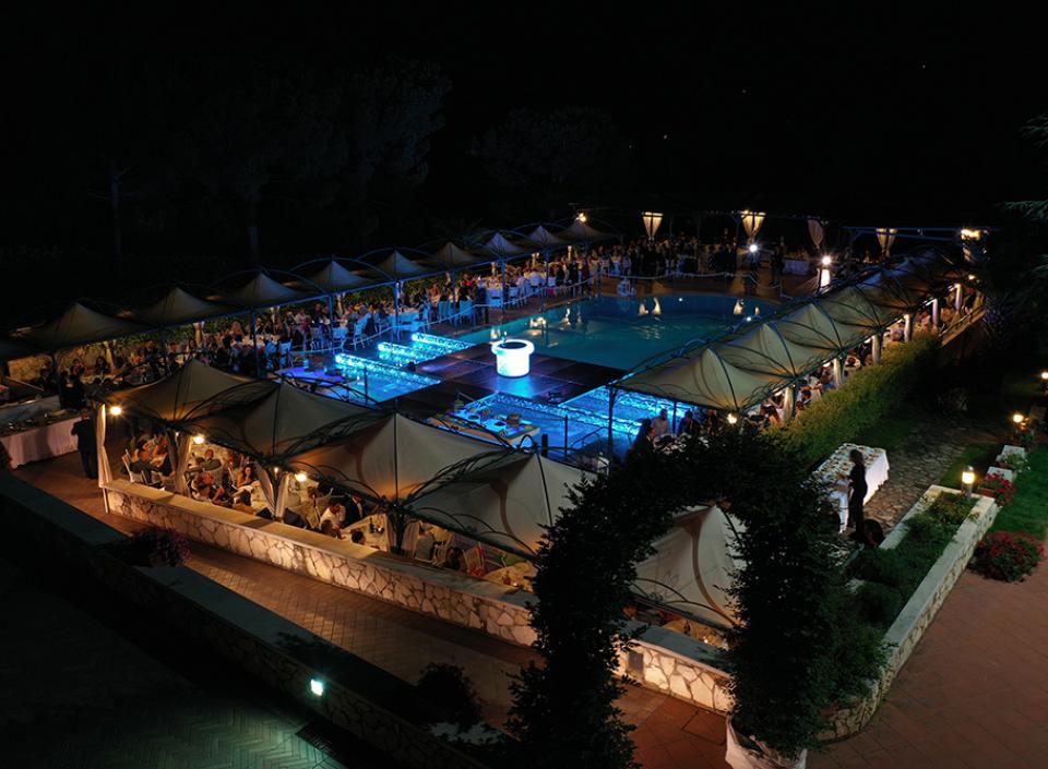 Festa notturna elegante, piscina illuminata, ospiti sotto tende, atmosfera magica e raffinata.