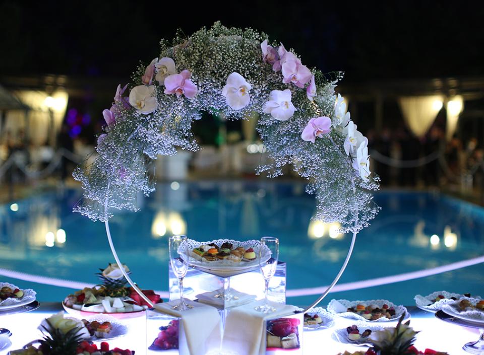 Tavola elegante con fiori sopraelevati, piatti raffinati, atmosfera serale intorno a una piscina.