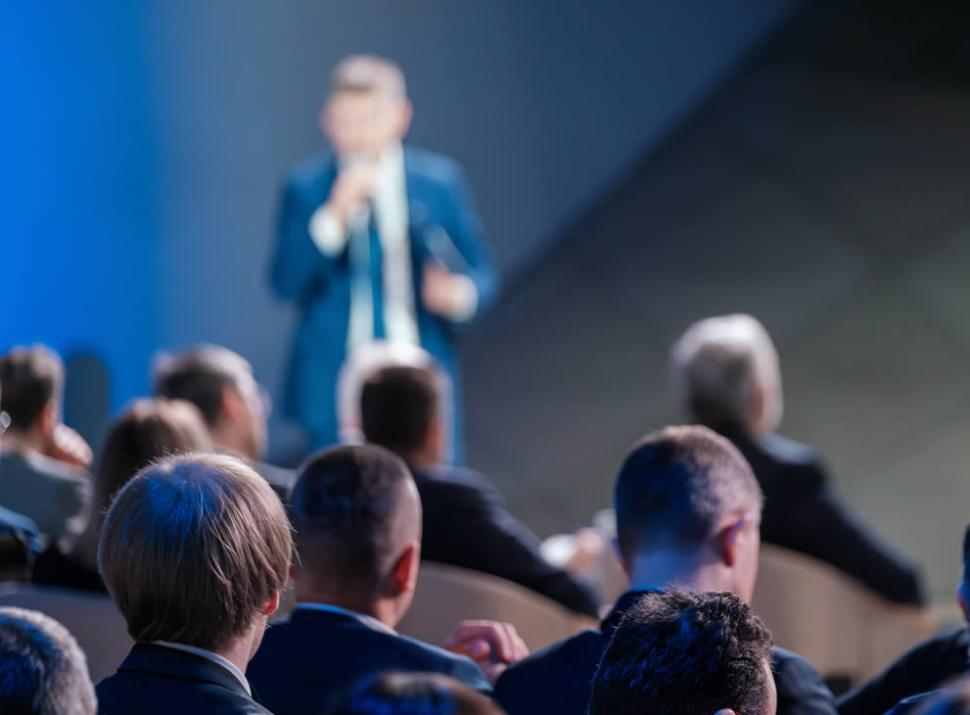 Conferenza aziendale con relatore e pubblico attento in abiti formali.
